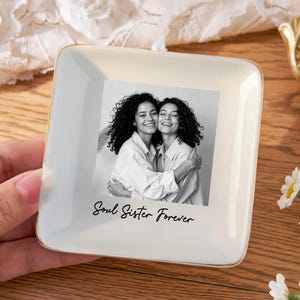 May include: Square, off-white ceramic trinket dish with a gold rim. The dish features a black and white photo of two women embracing, with the text "Soul Sister Forever" below the photo. A hand is holding the dish.