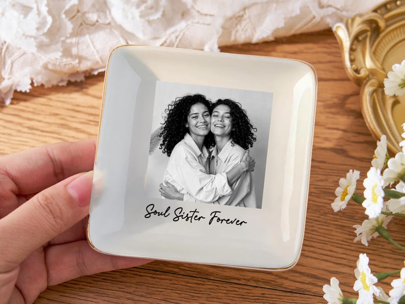 May include: Square, off-white ceramic trinket dish with a gold rim. The dish features a black and white photo of two women embracing, with the text "Soul Sister Forever" below the photo. A hand is holding the dish.