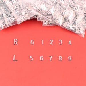 May include: Silver metal letters and numbers are displayed on a red background. The letters R and L, along with the numbers 0 through 9, are visible. Several bags of additional letters are in the background.