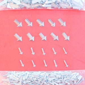 Könnte beinhalten: Silberfarbene, pfeilförmige Metallverzierungen auf rotem Hintergrund angeordnet. Die Pfeile zeigen nach unten und sind in verschiedenen Größen vorhanden. Ein durchsichtiger Plastikbeutel enthält weitere Verzierungen.