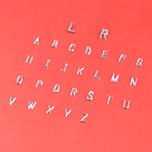 May include: Silver metal alphabet letters scattered on a red background. The letters are in a sans-serif font and are evenly spaced. The letters are small and shiny, and the background is a solid, bright red.