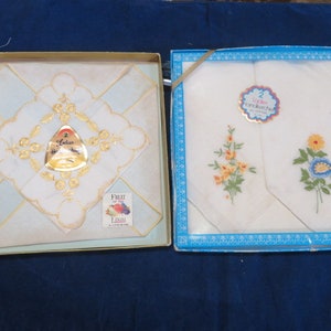 May include: Two boxes of vintage handkerchiefs. One box features a handkerchief with gold embroidery and a gold label. The other box contains two handkerchiefs with floral embroidery. The boxes are on a dark blue surface.