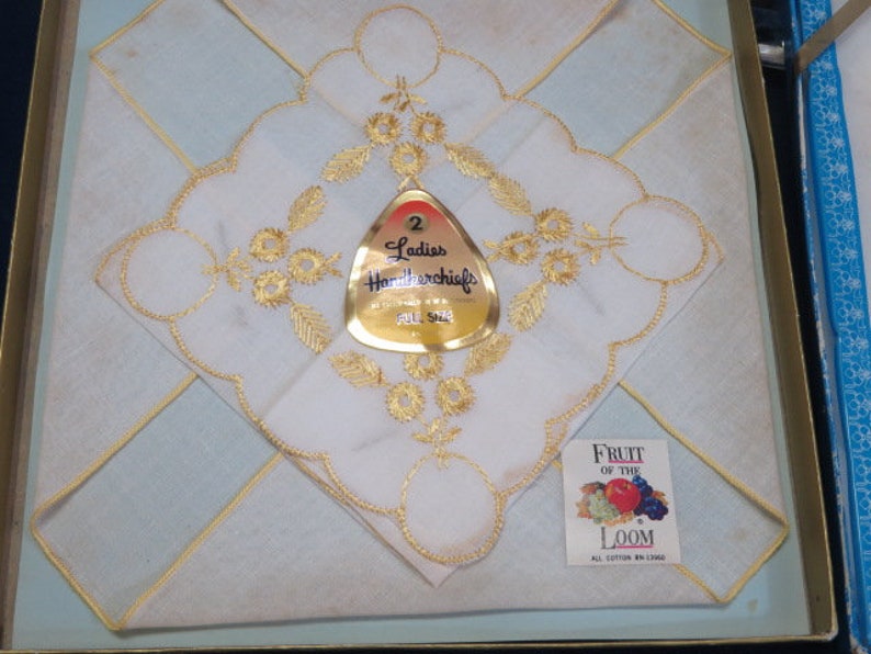 May include: A set of two white handkerchiefs with gold embroidered floral designs, presented in a box. The handkerchiefs feature intricate gold detailing and are labeled "Ladies Handkerchiefs". A small tag reads "Fruit of the Loom".