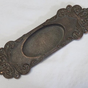 Peut inclure: Tirette de tiroir ornée de couleur bronze avec un centre ovale encastré. La plaque rectangulaire présente des volutes détaillées et des ornements décoratifs aux extrémités. Une pièce de quincaillerie de style vintage.