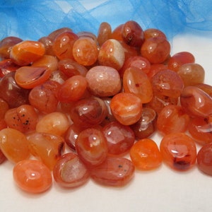 May include: A collection of carnelian tumbled stones in various shades of orange and red. The stones are smooth and polished, with a natural, earthy look.