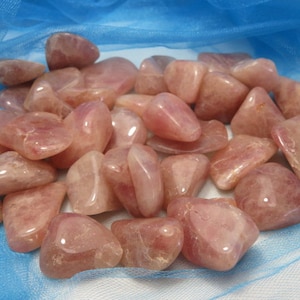 May include: A collection of tumbled rose quartz stones in various sizes and shapes. The stones are a pale pink color and have a smooth, polished finish.