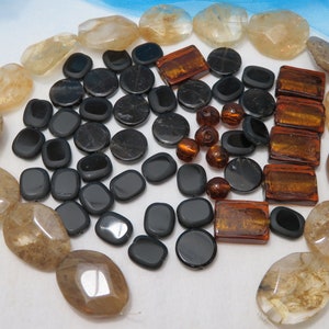 May include: A collection of black, brown, and clear glass beads. The beads are of various shapes and sizes, including round, oval, and rectangular. Some of the beads have a textured surface.