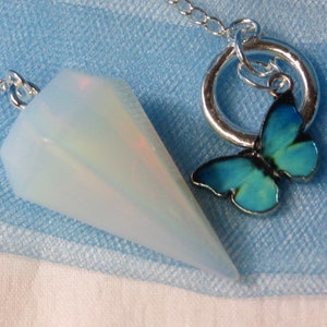 May include: A white opal pendulum with a silver chain and a blue butterfly charm. The pendulum is hanging from a silver ring.