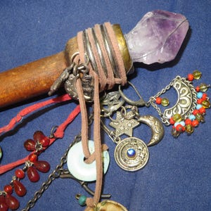 May include: A handcrafted wooden wand with a large, raw amethyst crystal at the tip. The wand is adorned with silver charms, leather cord, and beaded accents in red, turquoise, and amber. A unique, mystical item.