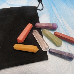 May include: A black velvet drawstring pouch accompanied by seven polished, hexagonal crystal wands in various colors, including clear, yellow, orange, red, green, purple, and dark blue, arranged on a light blue fabric surface.