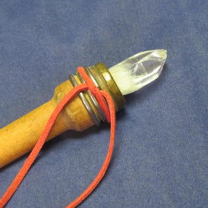May include: A handcrafted wand featuring a clear crystal point, set in a brass-colored metal casing. The wand's handle is made of light-colored wood, with a red cord wrapped around the metal. The background is a solid blue fabric.