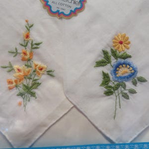 May include: Two white cotton handkerchiefs with embroidered floral designs. One features orange and yellow flowers with green leaves, the other has a blue and yellow flower with green leaves. The tag reads "ALL COTTON".