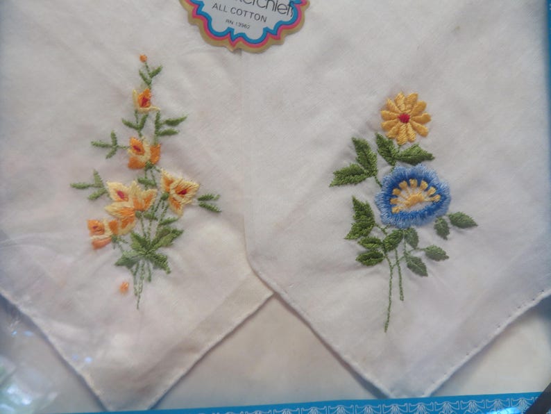 May include: Two white cotton handkerchiefs with embroidered floral designs. One features orange and yellow flowers with green leaves, the other has a blue and yellow flower with green leaves. The tag reads "ALL COTTON".