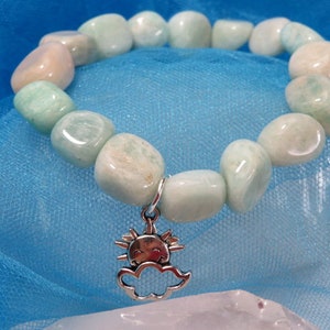May include: A beaded bracelet featuring light green and cream-colored stones. A silver charm of a smiling sun inside a cloud hangs from the bracelet. The bracelet is displayed on a blue fabric background.