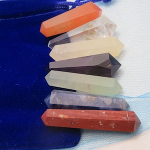 May include: A collection of seven polished crystal wands in various colors, including orange, clear, yellow, dark purple, green, blue, and red. The wands are arranged next to a dark blue velvet pouch.