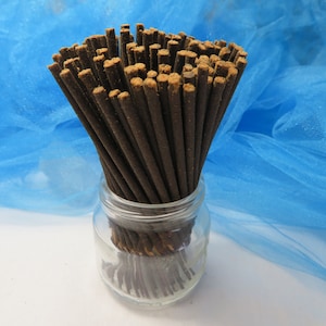 May include: A clear glass jar filled with numerous dark brown incense sticks. The incense sticks have a lighter brown tip. The jar sits on a white surface against a backdrop of blue fabric.