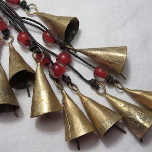 May include: A decorative hanging ornament featuring multiple cone-shaped brass bells suspended from dark brown leather cords. The design includes several translucent red glass beads interspersed along the cords, connecting the metallic bells.