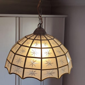 Puede incluir: Una lámpara colgante de estilo vintage con un diseño de estrella. La lámpara presenta una marquesina y una cadena de color latón, que sostienen una pantalla abovedada con un diseño de rejilla. Cada panel de la pantalla tiene un patrón de estrella. La luz cuelga del techo.