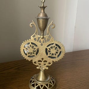 May include: A vintage brass decorative object with intricate filigree and floral designs. The object features a central vase-like structure, flanked by two circular elements with star-shaped patterns. The base has a geometric design. The object is likely a decorative piece.