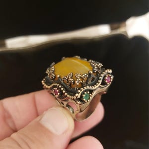 Può includere: Primo piano di un anello con una grande pietra preziosa gialla ovale. L'anello ha una montatura in metallo scuro ornata con filigrana e piccole pietre di accento rosse e verdi. L'anello è tenuto da una mano.