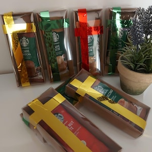 Może przedstawiać: Sześć małych pudełek prezentowych owiniętych w brązowy papier ze złotą wstążką. Każde pudełko zawiera inny smak kawy. Pudełka są ułożone w rzędzie na białej powierzchni.