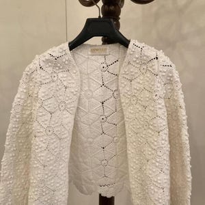 Pode incluir: Uma jaqueta de crochê branca com frente aberta e um padrão texturizado semelhante a flores. A jaqueta está pendurada em um cabide preto. O nome da marca "ASPROULIS" é visível no interior da jaqueta.