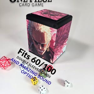 May include: A black and pink card storage box featuring a character from the One Piece anime. The box has the text "ONE PIECE CARD GAME" and "Fits 60/100 DOUBLE SLEEVED CARDS AND AMAZING SOUND OPTION!" Several dice are scattered around the box.