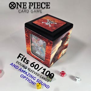 Op de afbeelding: Een zwarte en oranje One Piece kaartspelbox met de tekst "ONE PIECE CARD GAME" bovenaan. De doos heeft een venster met een kaartafbeelding en de tekst "Fits 60/100 Double Sleeved Cards and Amazing Sound Option!"
