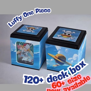 Può includere: Due scatole blu per mazzi con coperchi neri, con illustrazioni e testo di "Luffy One Piece". Ogni scatola ha un design a teschio e ossa incrociate sulla parte superiore. È visibile anche il testo "120+ deck box" e "60+ size now available".