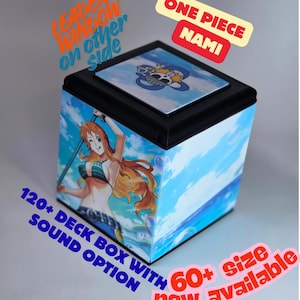 Op de afbeelding: Zwarte deckbox met het personage Nami uit One Piece, met een blauwe lucht en oceaan achtergrond. De doos heeft de tekst "ONE PIECE NAMI" en "120+ DECK BOX WITH SOUND OPTION". Verkrijgbaar in meer dan 60 maten.