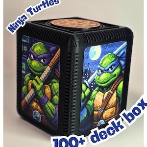 May include: Black deck box featuring pixelated images of Teenage Mutant Ninja Turtles. The box has a pizza-themed top and the text "Ninja Turtles" and "100+ deck box". The box is designed to hold cards.