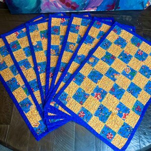 May include: A set of six quilted placemats. Each placemat features a checkerboard pattern of gold and blue squares, with floral accents. The edges are trimmed with a vibrant blue border. The placemats are arranged in a fan shape.