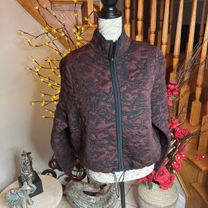 Puede incluir: Chaqueta acolchada con estampado burdeos y negro, expuesta sobre un maniquí. La chaqueta tiene cuello alto, cremallera completa y dobladillo con cordón. El fondo incluye una escalera y elementos decorativos.