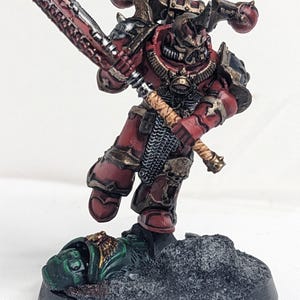 May include: A red and gold painted miniature of a space marine wielding a chain sword and standing over a fallen enemy. The miniature is posed in a dynamic action pose and is set on a black base with a gray and green textured surface.