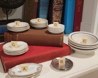 May include: A collection of small ceramic and metal dishes stacked in groups, each topped with a lettered tile. The items include white plates with gold rims, floral patterns, and blue bands, arranged on and around two large hardcover books.
