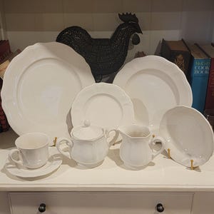Puede incluir: Un conjunto de vajilla de cerámica blanca que incluye dos platos grandes, un plato pequeño, un cuenco, una taza de té con platillo, un azucarero con tapa y una jarra de leche. Cada pieza presenta un borde festoneado y un acabado liso y brillante.