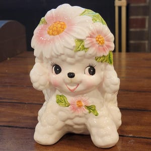 May include: A white ceramic poodle figurine featuring a glossy finish, large black eyes, and a pink nose. The poodle is adorned with sculpted pink daisies with yellow centres and green leaves on its head and chest, showcasing detailed fur texture.