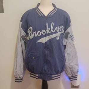 Könnte beinhalten: Eine blaue Reißverschluss-Collegejacke mit hellblauen Nadelstreifen und hellblauen Ärmeln. Auf der Vorderseite steht das Wort Brooklyn in weißer Schreibschrift. Sie hat eine kleine Brusttasche mit Knopf sowie gestreifte Rippbündchen.
