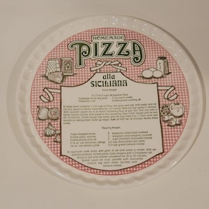 May include: A round white pizza pan with a scalloped edge featuring a pink and white checkered border. The center displays the text Homemade Pizza alla Siciliana, along with illustrations of ingredients and a detailed crust and topping recipe.