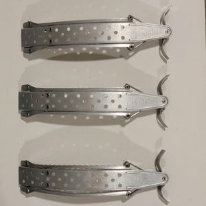 May include: Three identical silver-toned metal hair barrettes arranged vertically. Each barrette features a long, perforated rectangular body with a curved, spring-loaded clasp mechanism at one end and a hinged closure at the other end.