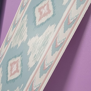 Peut inclure: Une bande de papier peint présentant un motif ikat répétitif dans des tons doux de bleu cendré, rose pâle et crème. Le design se compose de formes en losange et de lignes verticales texturées bordées de fines rayures droites couleur crème.