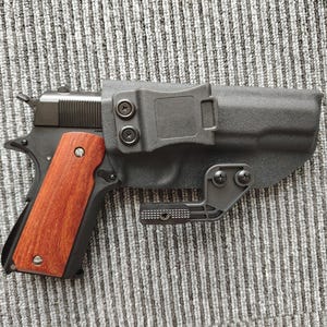 May include: A black handgun with a wooden grip is holstered in a black leather holster with a metal clip.