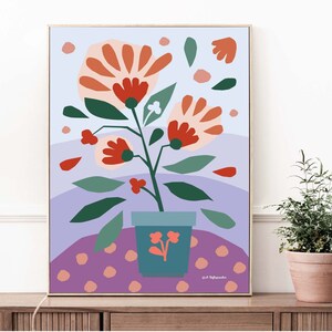 Op de afbeelding: Een ingelijste kunstdruk met een gestileerde plant met grote oranje en rode bloemen, groene bladeren en een blauwgroene pot. De achtergrond bevat tinten paars en een stippenpatroon. Het kunstwerk wordt getoond op een houten oppervlak.