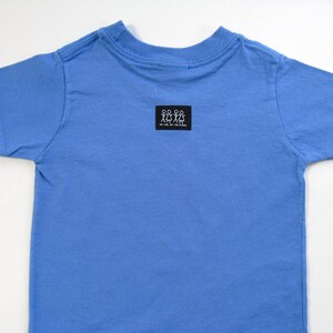 Pode incluir: Uma camiseta azul clara de manga curta. A camisa tem gola redonda e um pequeno patch retangular preto perto da gola com um gr&aacute;fico branco de figuras de palito. O texto no patch &eacute; ileg&iacute;vel.