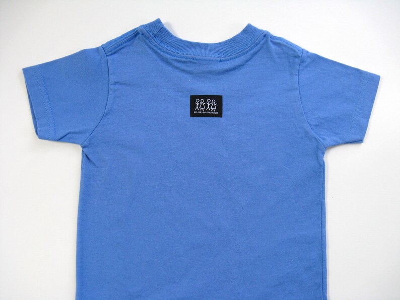 Pode incluir: Uma camiseta azul clara de manga curta. A camisa tem gola redonda e um pequeno patch retangular preto perto da gola com um gr&aacute;fico branco de figuras de palito. O texto no patch &eacute; ileg&iacute;vel.