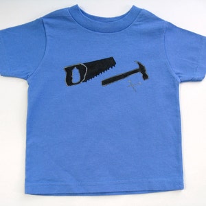 Pode incluir: Uma camiseta azul clara com um desenho de serrote e martelo pretos. As ferramentas s&atilde;o delineadas em preto e parecem desenhadas &agrave; m&atilde;o. A camiseta &eacute; feita de material macio e tem gola redonda.