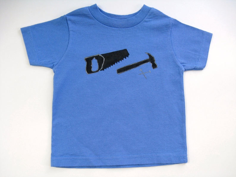 Pode incluir: Uma camiseta azul clara com um desenho de serrote e martelo pretos. As ferramentas s&atilde;o delineadas em preto e parecem desenhadas &agrave; m&atilde;o. A camiseta &eacute; feita de material macio e tem gola redonda.