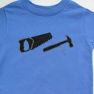 Pode incluir: Camiseta azul claro com um desenho preto de serrote, martelo e pregos. O serrote e o martelo t&ecirc;m um contorno mais claro. Uma camiseta infantil com ferramentas.