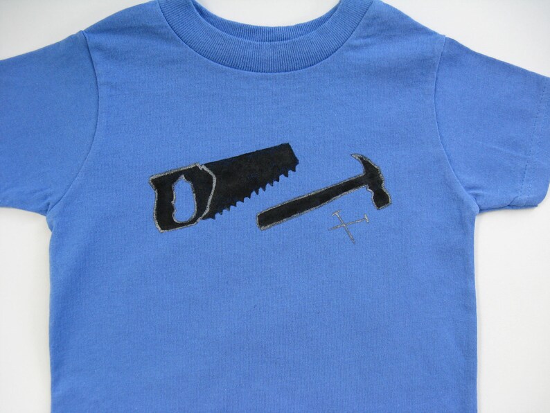 Pode incluir: Camiseta azul claro com um desenho preto de serrote, martelo e pregos. O serrote e o martelo t&ecirc;m um contorno mais claro. Uma camiseta infantil com ferramentas.