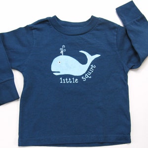 May include: A blue long-sleeved shirt with a light blue whale graphic and the text "little squirt" on the front.